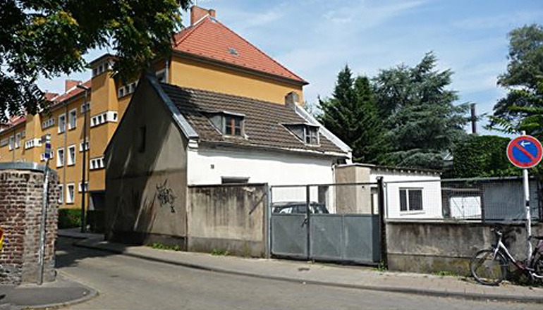 Bild von der Begehung des Herzhäuschens in Köln Bickendorf vor dem Abriss: Blick von der Straße aus. Das Grundstück wird von einer Mauer und einem Tor begrenzt.